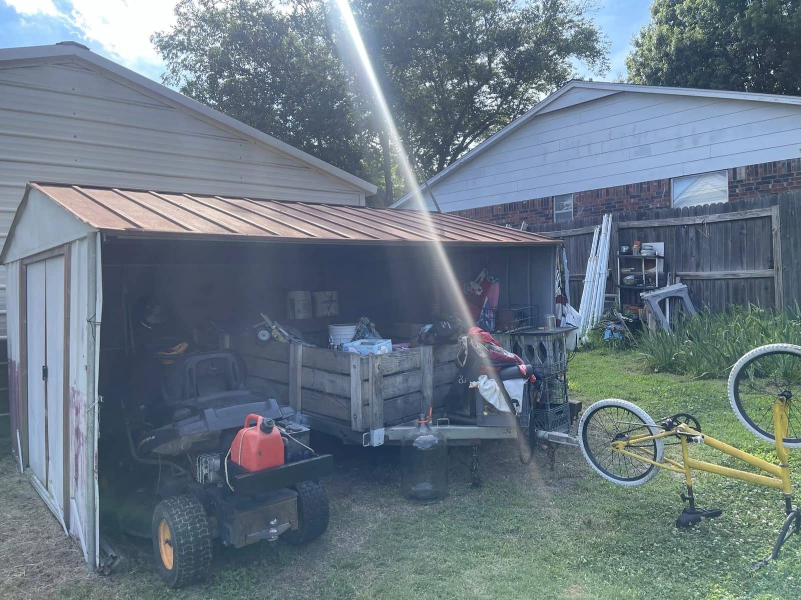 A cluttered backyard storage shed in Tulsa before clearing, packed with a riding mower, a junk-filled utility trailer, paint cans and shelving, with a bike lying on the grass outside