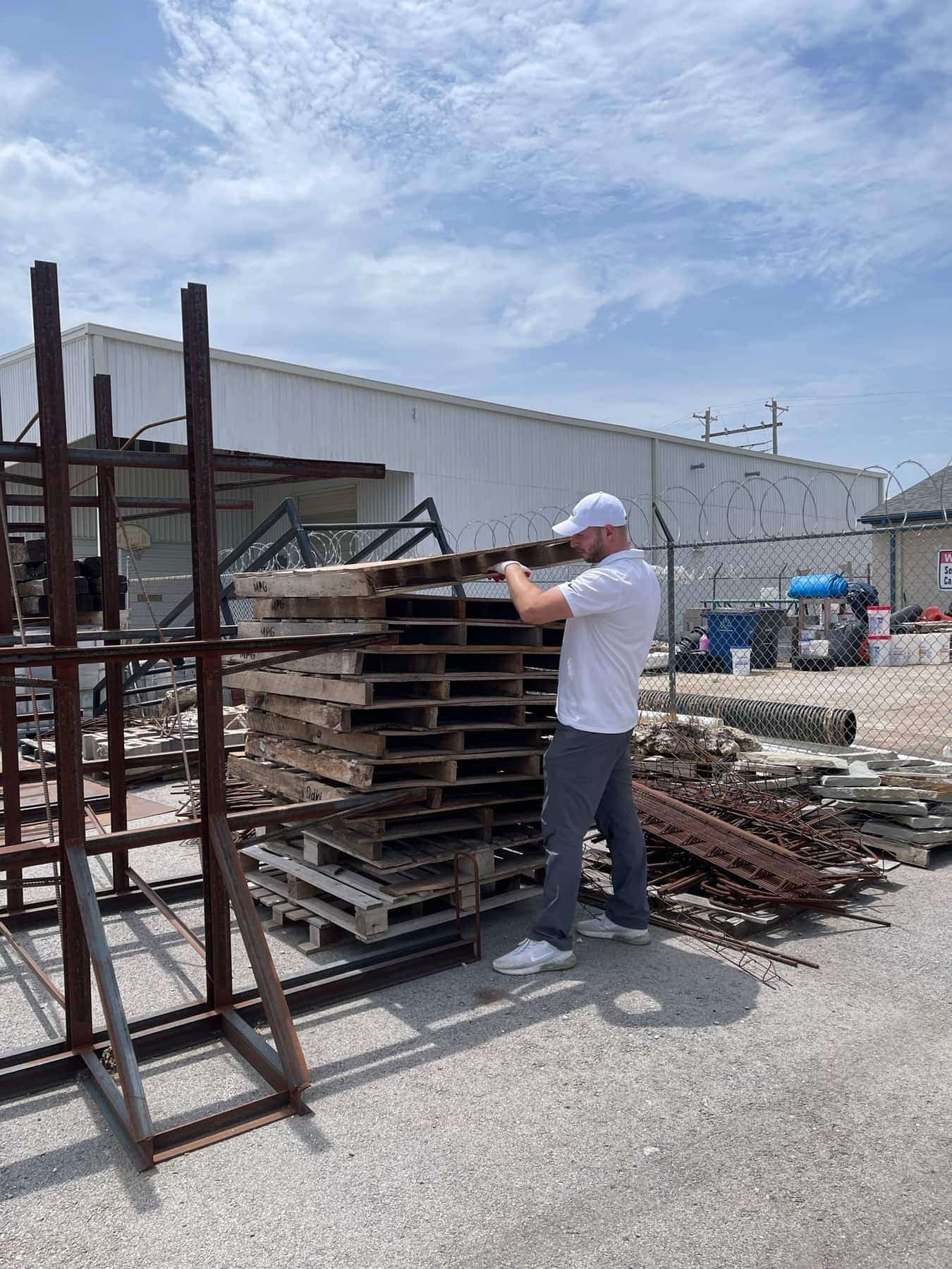 Victor lifting a wooden pallet off a stack at a Tulsa scrap yard, with steel racking and rebar behind him