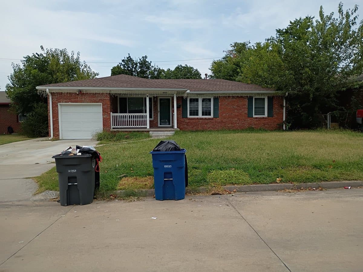 The same red brick Tulsa ranch house photographed from the street with the verge cleared — nothing left on the grass, only the homeowner’s grey trash cart and blue recycling cart standing at the curb with bags on top