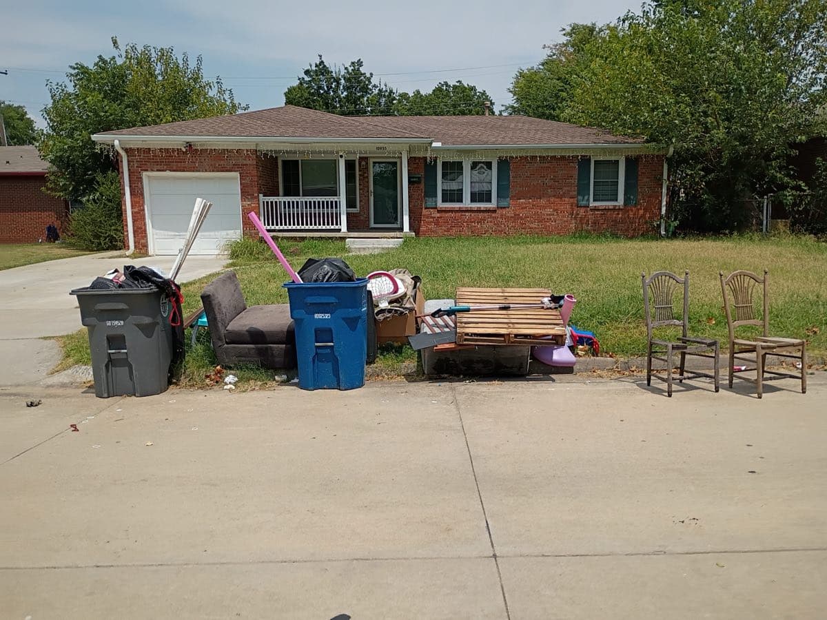 Household junk piled on the grass verge outside a red brick Tulsa ranch house — a brown fabric loveseat, a wooden pallet, two wooden ladder-back dining chairs, a cardboard box, loose boards and a black bin bag — with the grey trash cart and blue recycling cart standing beside it
