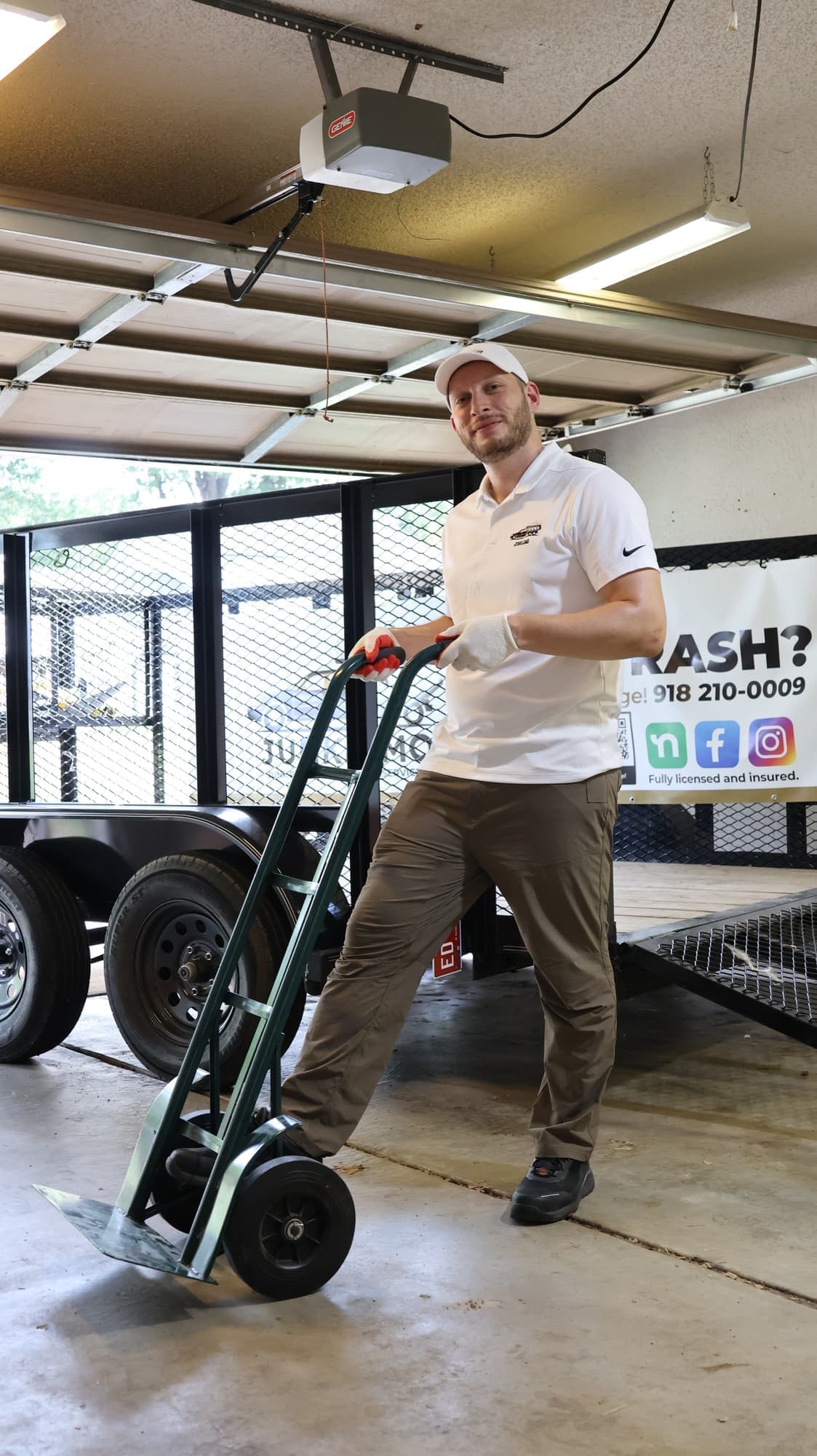 Victor wheeling a hand truck past the open trailer in the JunkLimo garage, in front of a banner showing the phone number (918) 210-0009