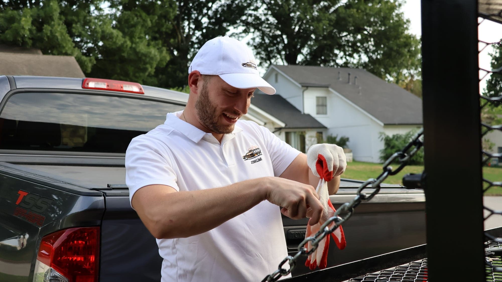 Victor pulling on red and white work gloves at the open tailgate of his truck outside a Tulsa house